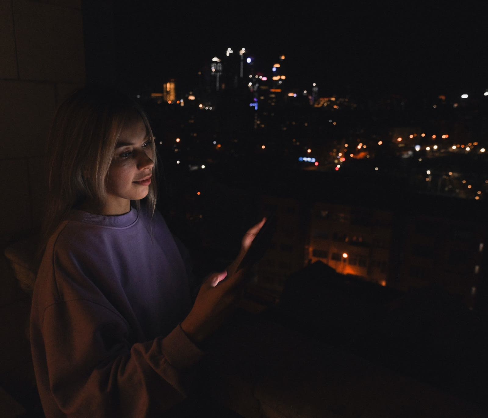 A young woman on a balcony at night, her face lit by her phone, the city spread out below her