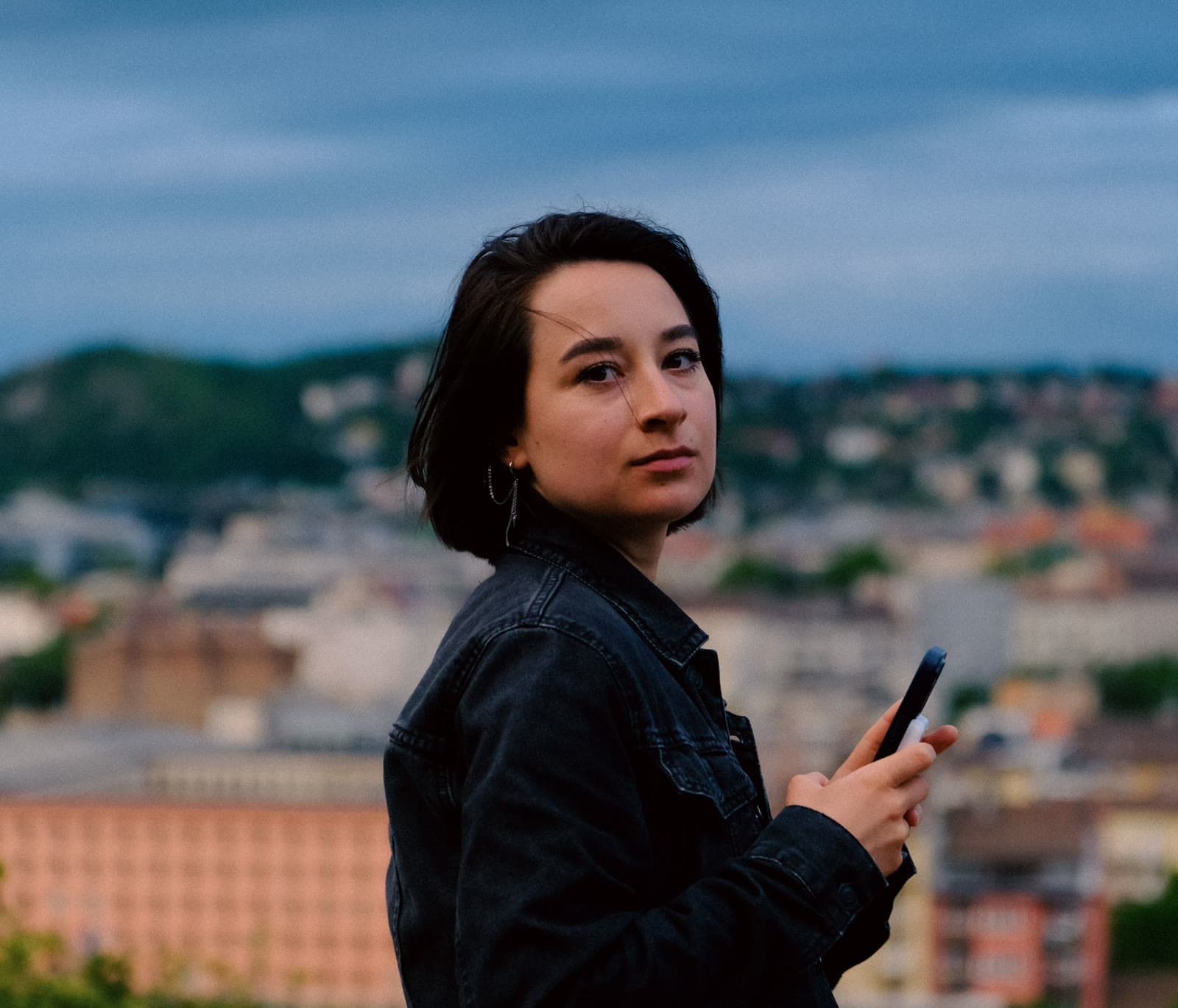 A young woman holding her phone, looking out across a city